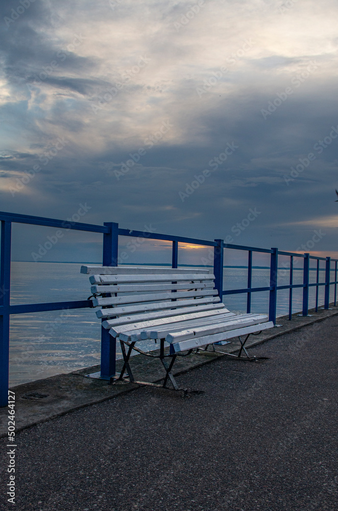An empty wooden bench standing on the pier on the Vistula Lagoon in Frombork, Poland against the setting sun; evening