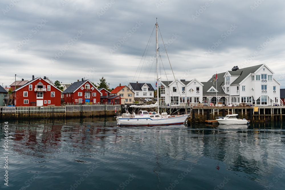 Norwegian seascape, cityscape of the town Henningsvaer, a small boat moves between peninsulas, sail boat, rocky coast with dramatic skies, classic view of Norwegian houses on the slopes Norwegian seascape, cityscape of the town Henningsvaer, a small boat moves between peninsulas, sail boat, rocky coast with dramatic skies, classic view of Norwegian houses on the slopes