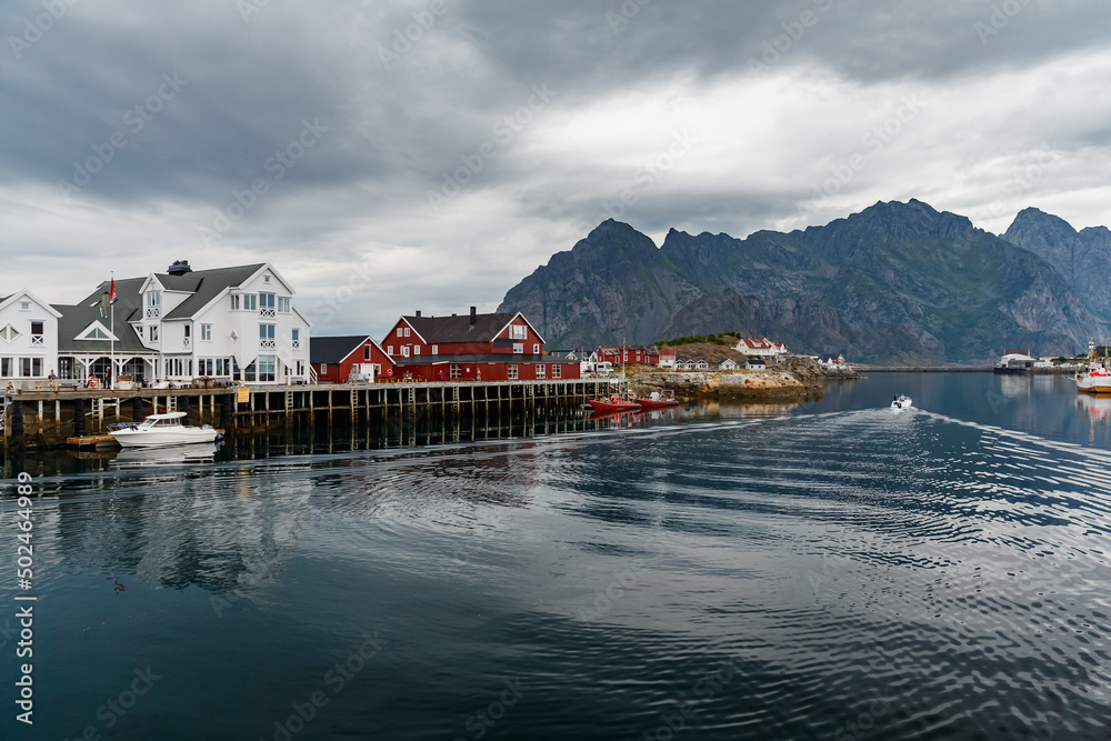 Norwegian seascape, cityscape of the town Henningsvaer, a small boat moves between peninsulas, sail boat, rocky coast with dramatic skies, classic view of Norwegian houses on the slopes