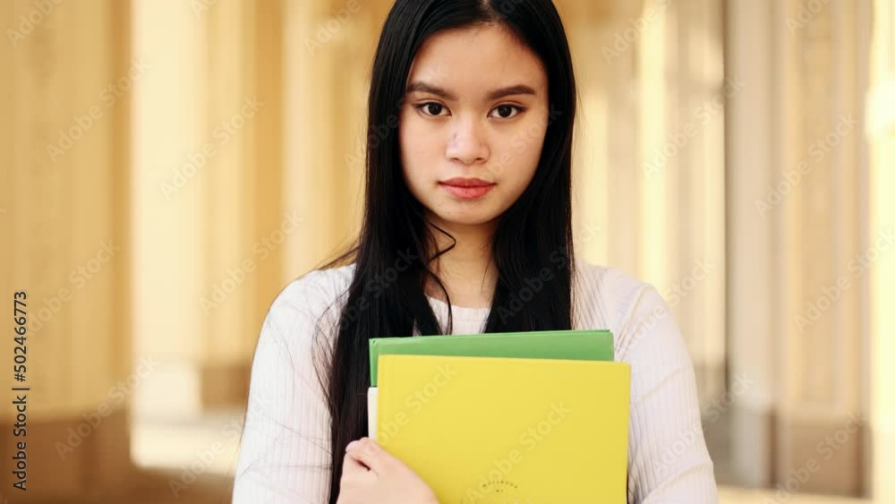 Serious asian student girl with books standing near the university building and looking at the camera. Portrait of attractive girl. Serious asian student girl with books standing near the university building and looking at the camera. Portrait of attractive girl.
