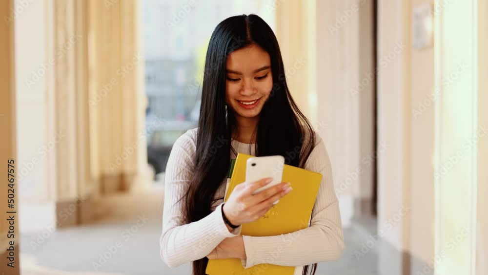 Portrait of attractive student young asian woman walking near the university building hold smartphone, watching social media. Smiling young woman texting on her phone. Portrait of attractive student young asian woman walking near the university building hold smartphone, watching social media. Smiling young woman texting on her phone.