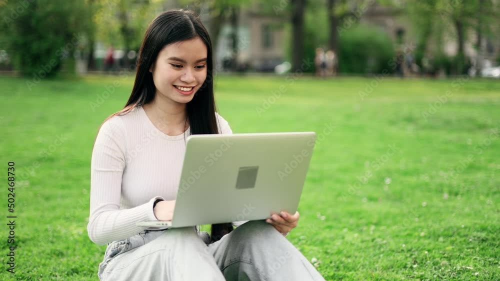 Dreamy attractive asian woman sitting on a grass in the park and talking on laptop by video chat. Student learning online. Dreamy attractive asian woman sitting on a grass in the park and talking on laptop by video chat. Student learning online.