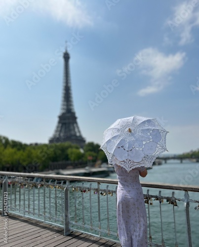 from the back we see a girl in a beautiful long dress in a retro style with an umbrella who walks towards the eiffel tower holding the dress with one hand. High quality photo
