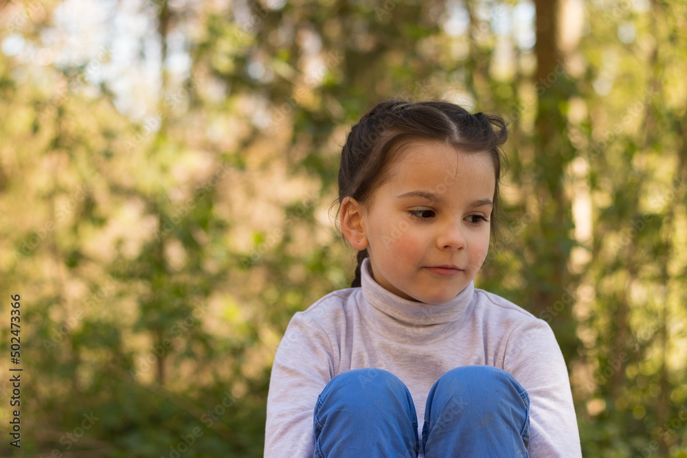 A small beautiful smiling schoolgirl on a natural background in a turtleneck sweater and jeans sits on the ground hugging her legs and looking away A small beautiful smiling schoolgirl on a natural background in a turtleneck sweater and jeans sits on the ground hugging her legs and looking away