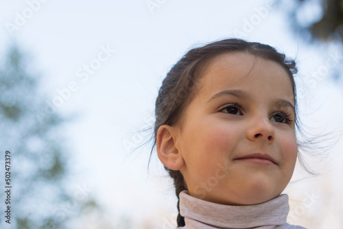 Close-up portrait against the blue sky of a beautiful first-grader girl in a gray turtleneck sweater