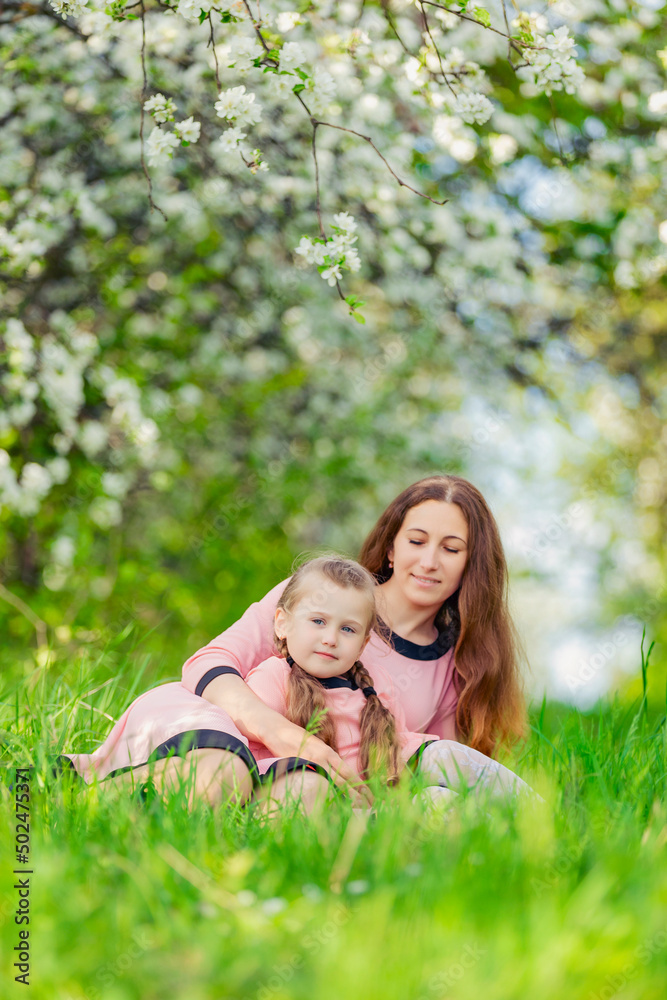 mother and daughter sit in the green grass against the backdrop of blooming apple trees mother and daughter sit in the green grass against the backdrop of blooming apple trees