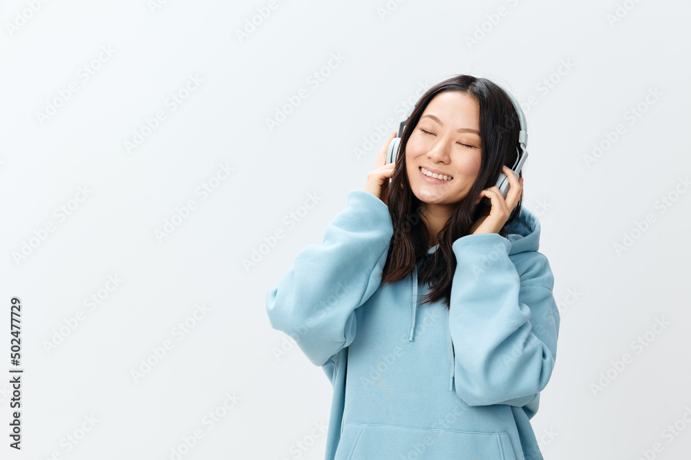 Overjoyed happy pretty Korean young female in blue hoodie touch headphones relax closing eyes posing isolated on over white studio background. The best offer for ad. Music App Platform Ad concept Overjoyed happy pretty Korean young female in blue hoodie touch headphones relax closing eyes posing isolated on over white studio background. The best offer for ad. Music App Platform Ad concept