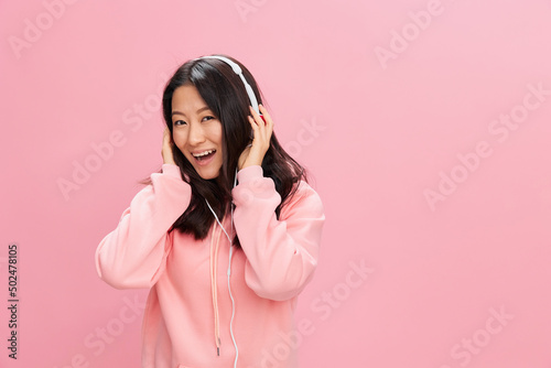 Enjoying Asian student young lady in pink hoodie sweatshirt with cute headphones smile touch head posing isolated on over pink studio background. Good offer. Sound streaming platform ad concept