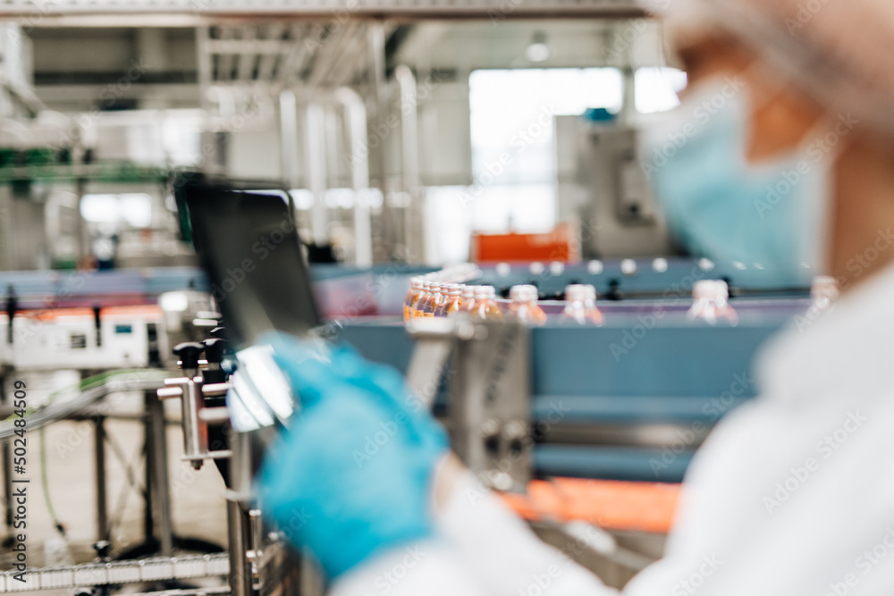 Beautiful female worker in workwear, with disposable protective cap and with protective mask on her face working in bottling factory. Inspection quality control. Beautiful female worker in workwear, with disposable protective cap and with protective mask on her face working in bottling factory. Inspection quality control.