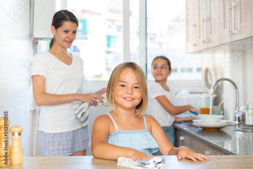 Little smiling girl cleaning kitchen surfaces at home while her mother and sister washing dish