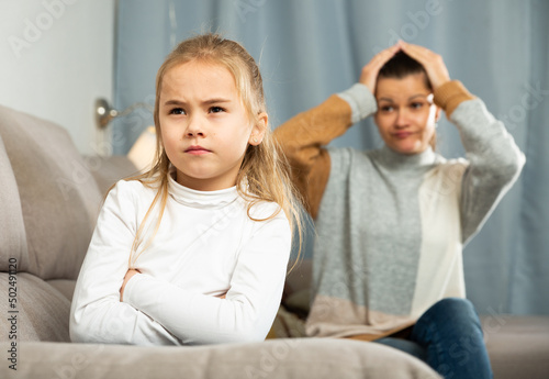 Frustrated small girl sitting at sofa, having conflict with mother at home interior