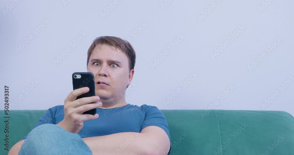 Young man with bored look uses smartphone, but suddenly sees shocking news. He opens his eyes and mouth wide in surprise. Woman and dachshund dog come to check what happened Young man with bored look uses smartphone, but suddenly sees shocking news. He opens his eyes and mouth wide in surprise. Woman and dachshund dog come to check what happened