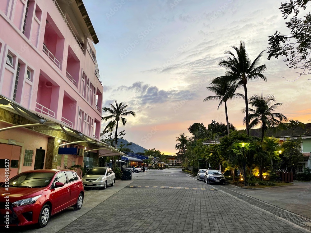 Sunset. Street of tropical town. Empty road paved with stones. Pink residential building. Pink beautiful glow of the sun on the sky. Cars parked along the side of the road at the curb. Several people