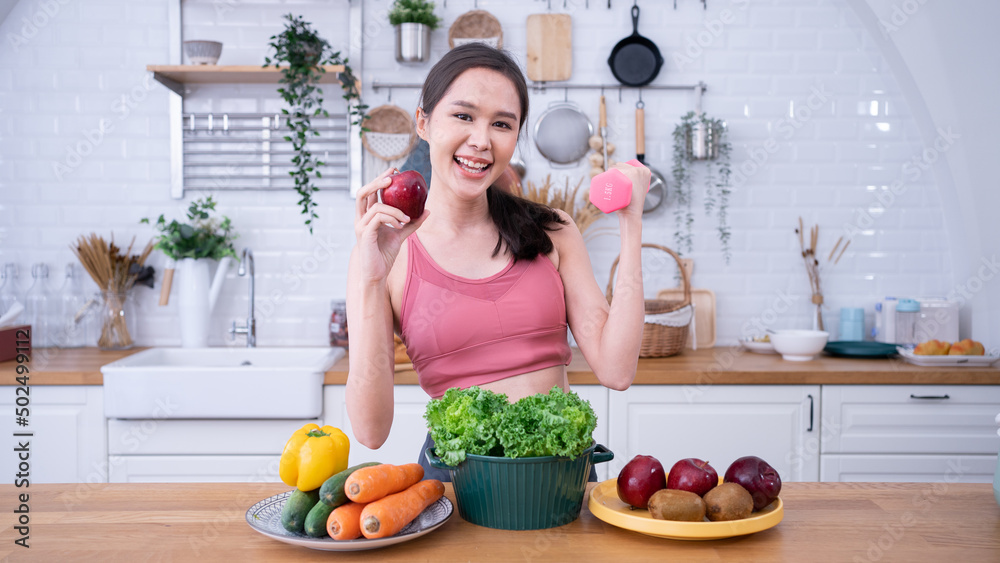 Healthy woman cooking organic vegetable salad in sportswear, yoga.Asian woman holding apples,dumbbells,vegetarian diet prepare food in  kitchen.
Food rich in vitamins, nourishing the body,happy,skin.