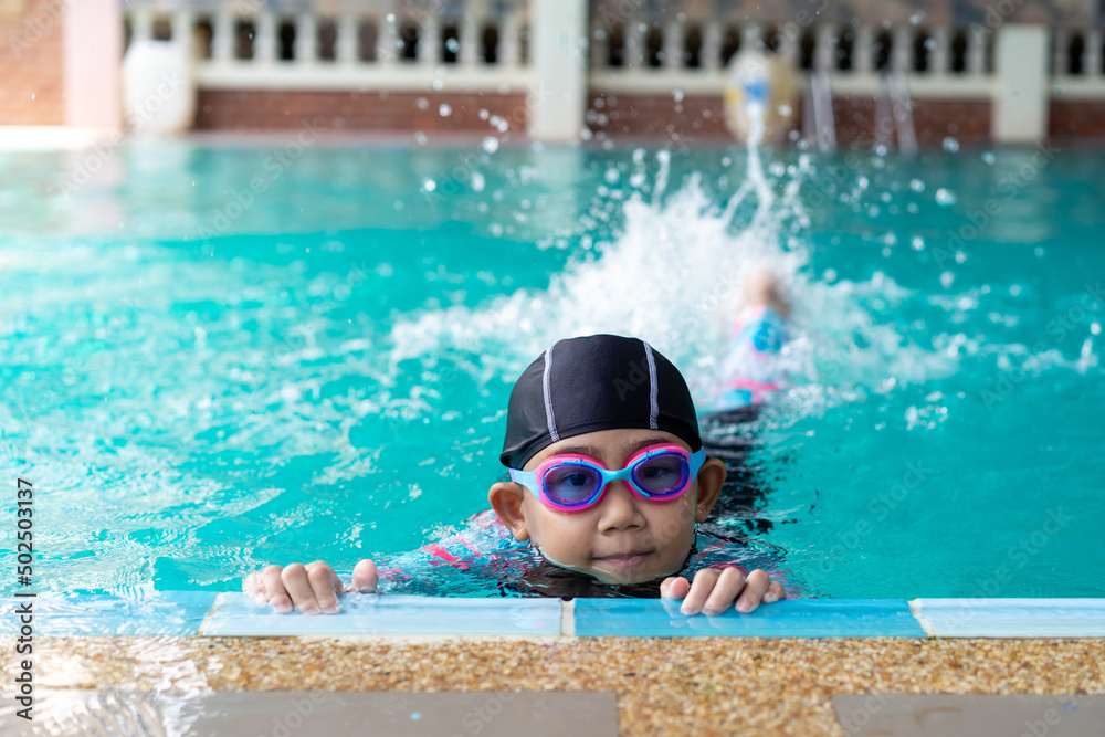 Asian child little girl wearing swimsuit and cap with goggles on edge swimming pool. Learn to swim idea concept