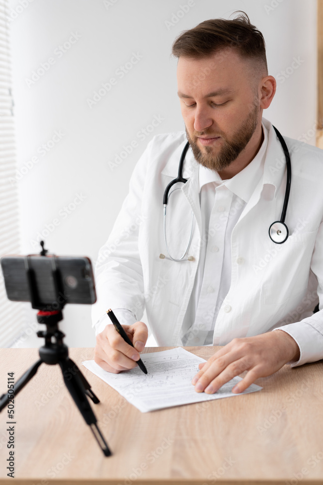 Caucasian young male doctor in a bathrobe with a stethoscope makes an online consultation using a smartphone. Caucasian young male doctor in a bathrobe with a stethoscope makes an online consultation using a smartphone.