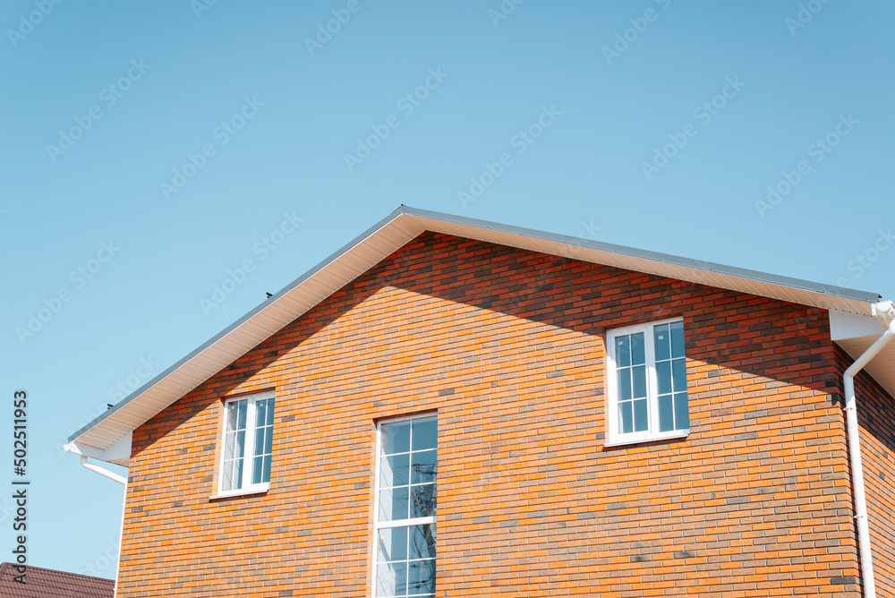 Exterior part of residential two-story house made of red brick with windows and roof against clear blue sky, outdoors