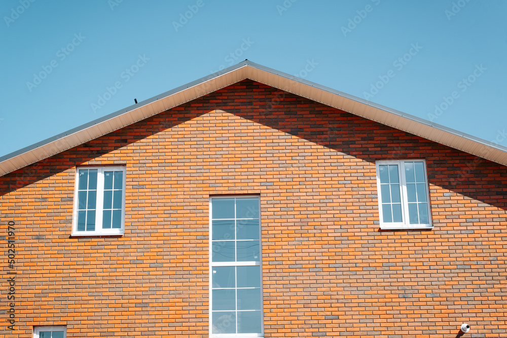 Exterior part of residential two-story house made of red brick with windows and roof against clear blue sky, outdoors. Front view, close-up