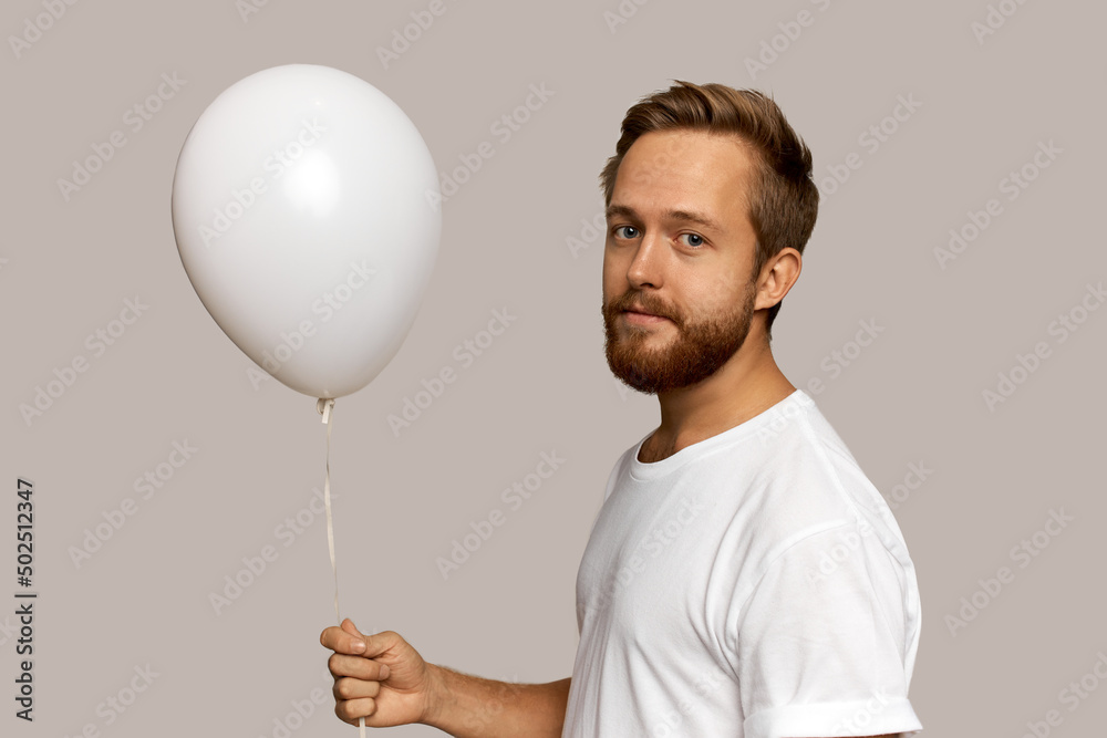 Cute red-headed unshaven guy standing against gray studio wall half-turned to camera with white air balloon with copy space for your advertisement in right hand, having calm face expression Cute red-headed unshaven guy standing against gray studio wall half-turned to camera with white air balloon with copy space for your advertisement in right hand, having calm face expression