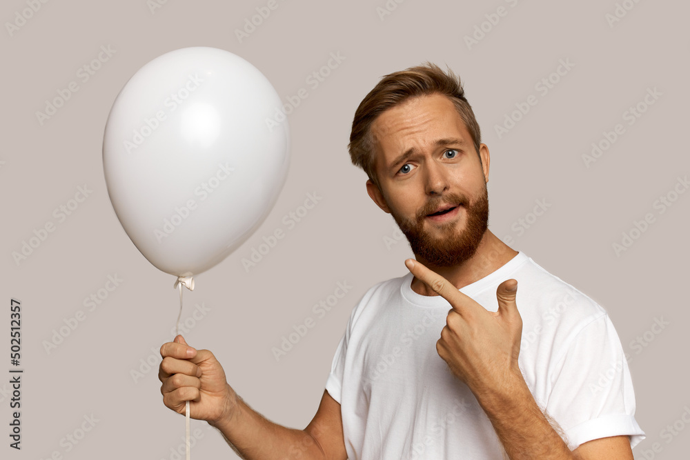 Funny-looking Caucasian red-headed man in sport shirt standing over gray background pointing at white inflatable helium balloon while holding it, having puzzled questioning facial expression Funny-looking Caucasian red-headed man in sport shirt standing over gray background pointing at white inflatable helium balloon while holding it, having puzzled questioning facial expression