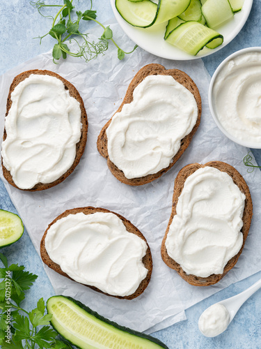 Open sandwiches, rye bread bruschetta with cream cheese spread (ricotta) and cucumber slices on white wax paper. Healthy delicious breakfast, lunch or snack food. Top view. Blue background.