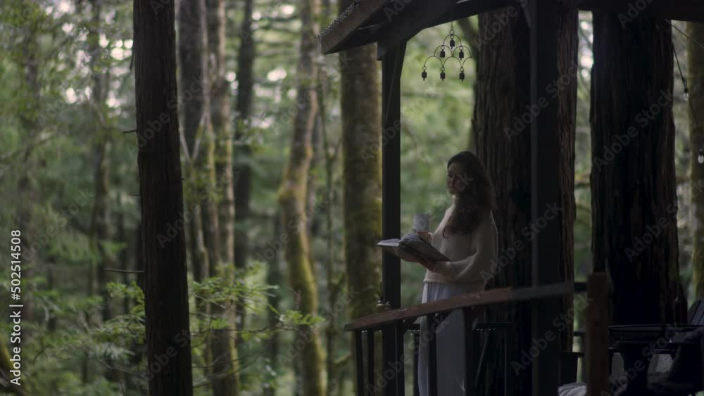 Young woman reading a book and drinking hot tea on a porch of a cozy cabin in the woods. Log cabin in the middle of woods sunny wooden bright mountain cabin rustic house architecture. Young woman reading a book and drinking hot tea on a porch of a cozy cabin in the woods. Log cabin in the middle of woods sunny wooden bright mountain cabin rustic house architecture.