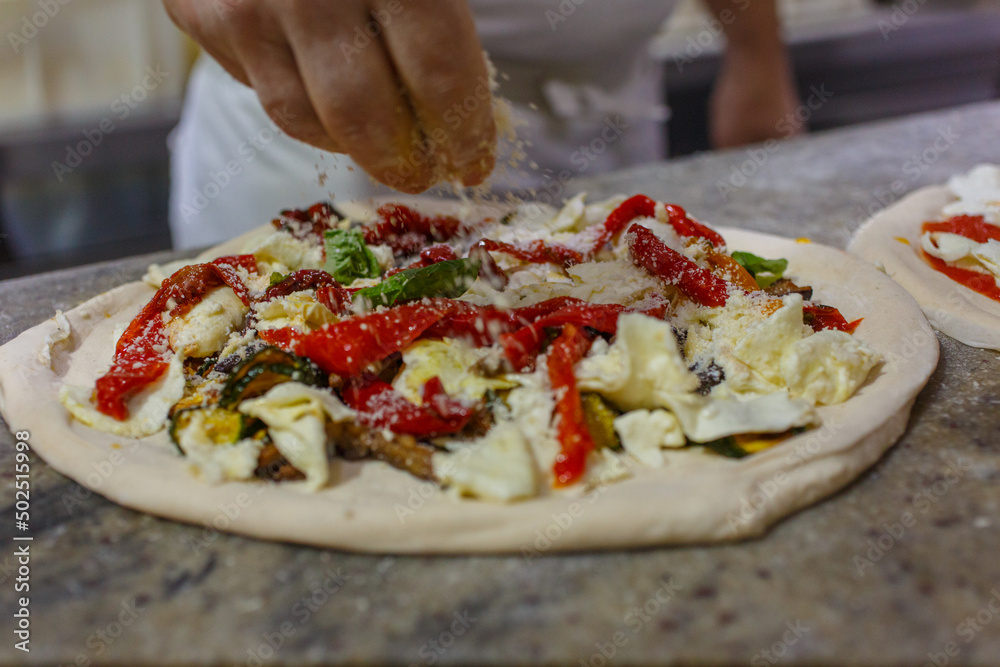 Pizzaiolo napoletano mentre mette del formaggio su una pizza napoletana pronta per essere infornata con peperoni, melanzane, zucchine e mozzarella di bufala