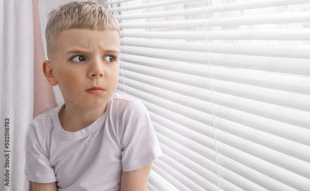 a sad little boy with blond hair in a white T-shirt sits alone at home at a window with blinds and looks sadly at the street. social concept a sad little boy with blond hair in a white T-shirt sits alone at home at a window with blinds and looks sadly at the street. social concept