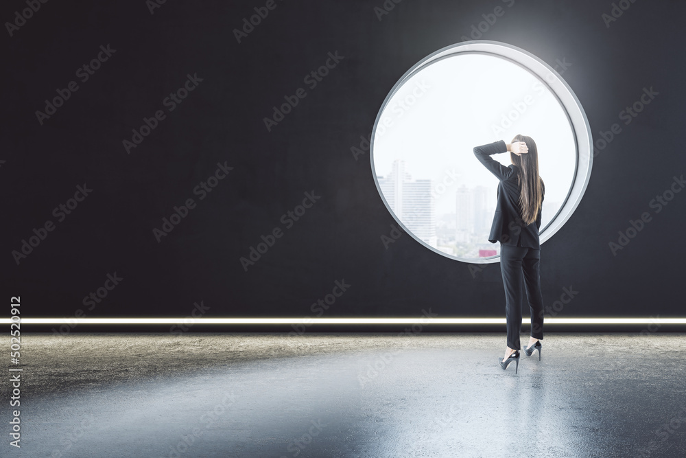 Obraz premium Back view of thoughtful young businesswoman standing in creative modern concrete interior with round window and city view. Mock up place on wall and daylight.