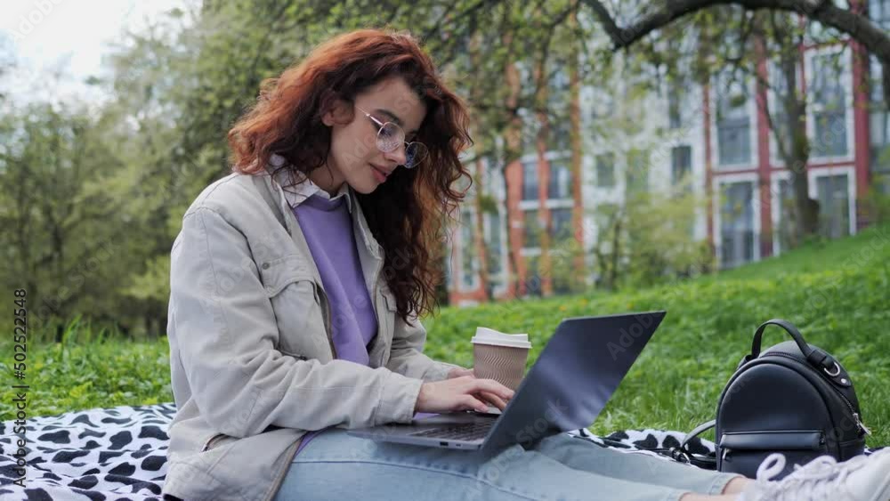 Attractive girl student is typing on the modern laptop holding coffee outdoors. Young woman with curly hair and glasses doing remotely job, working online at a laptop sitting on the grass, fresh air. Attractive girl student is typing on the modern laptop holding coffee outdoors. Young woman with curly hair and glasses doing remotely job, working online at a laptop sitting on the grass, fresh air.