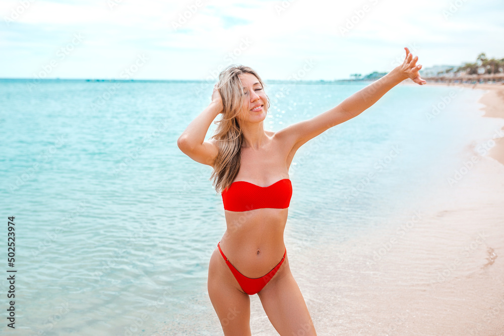 Hello. Young woman resting alone on the beach. Blonde girl in a red swimsuit walks and looks at the sea view of a tropical beach, travel and vacation on the ocean. Happy summer lifestyle on vacation