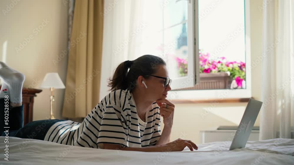Handheld cinematic shot of a young woman wearing glasses using her laptop while lying on a bed in a hotel room. Handheld cinematic shot of a young woman wearing glasses using her laptop while lying on a bed in a hotel room.