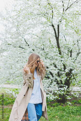 A young woman with blond hair enjoys a blooming spring garden. Travel, spring break. Fashionable style. A woman in sunglasses and a beige trench coat runs through a flowering park. Selection focus