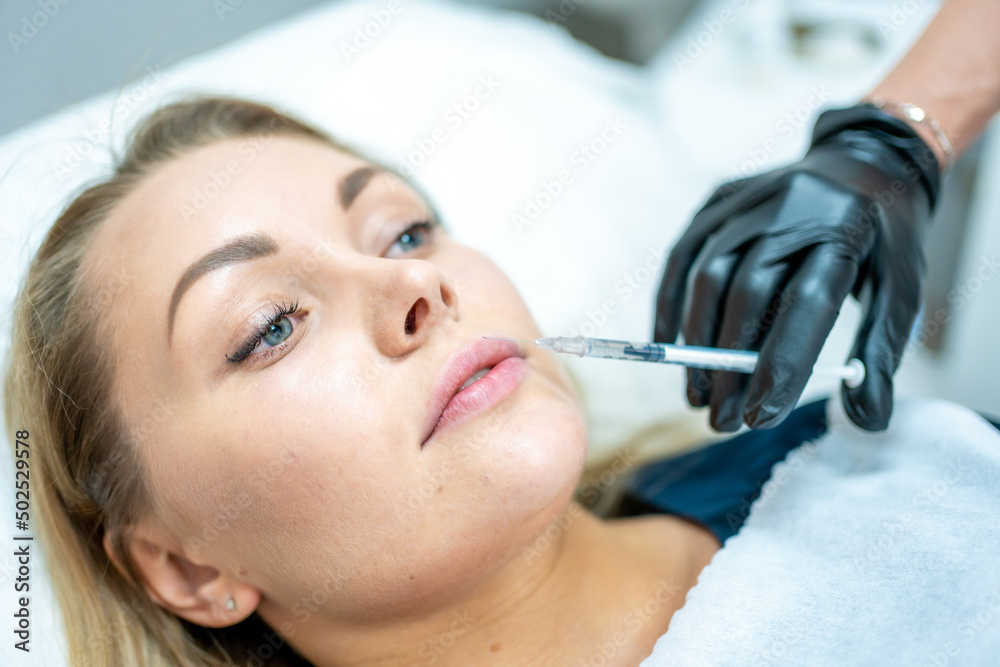 Close-up of woman face and hand in surgical glove holding syringe near her lips, ready to receive beauty treatment. Injection cosmetology, lips augmentation and correction concept.