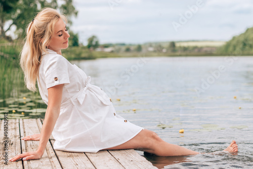 A young blonde pregnant woman in a white dress is sitting on a bridge by the river, with her feet in the water.Summer,pregnancy and prenatal care concept. © Tatyana
