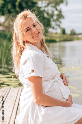 Tableau sur toile A young blonde pregnant woman in a white dress is sitting on a bridge by the river, with her feet in the water