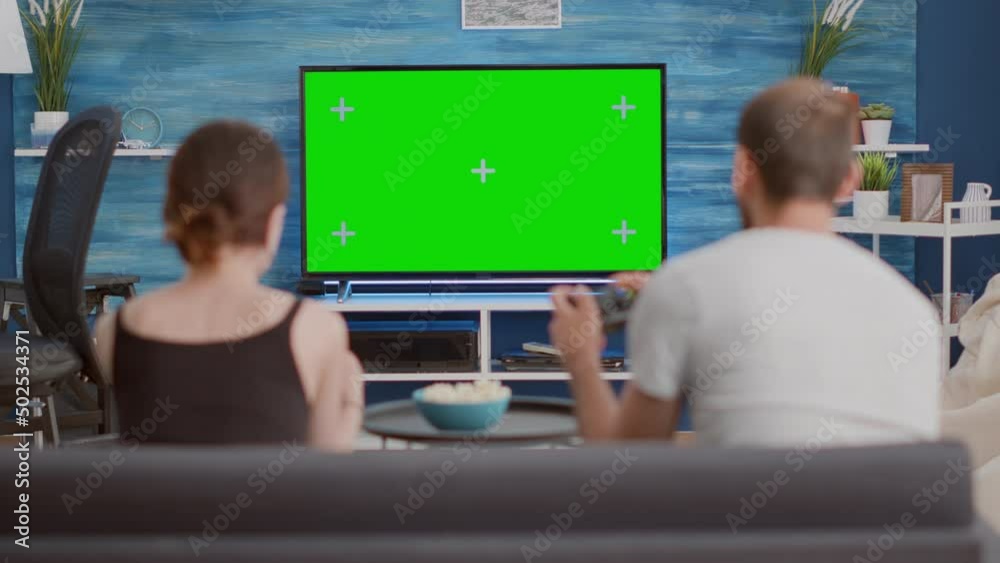 Couple holding controllers playing console online games on green screen tv sitting on couch in modern living room. Young woman and boyfriend spending free time gaming on chroma key mockup display.