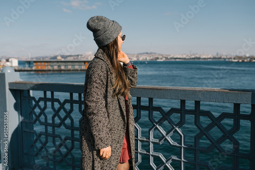 A girl with dark long hair in glasses, in gray coats and a hat looks at the seafront in Istanbul. A student travels during a pandemic. Walks in the autumn in the fresh air. Photo from the back, the