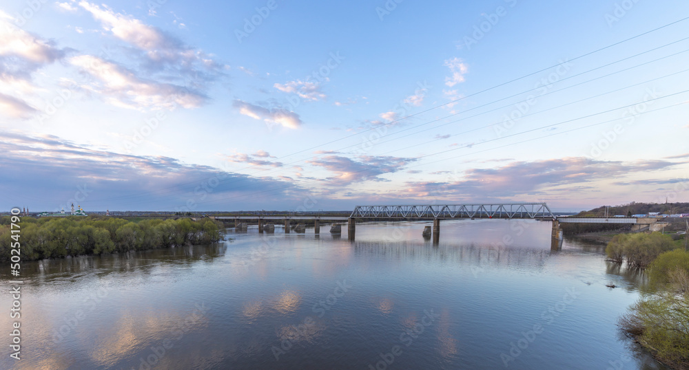 Evening landscape, sunset on the river. View of the industrial area of the railway bridge. Outskirts.