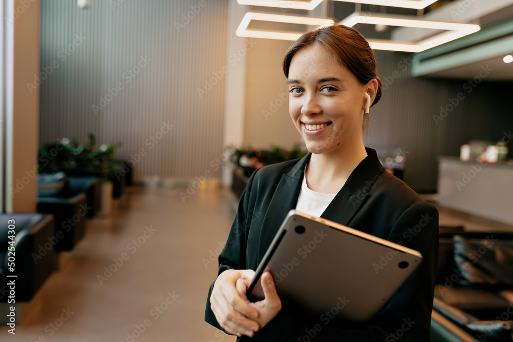 Smiling confident woman with collected hair wearing dark jacket and white t-shirt is holding laptop and smiling while working in office Smiling confident woman with collected hair wearing dark jacket and white t-shirt is holding laptop and smiling while working in office