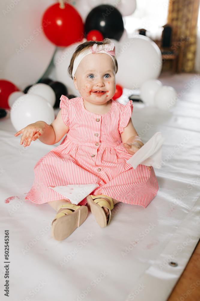 Fototapeta premium beautiful little one-year-old girl in a pink dress eats a lollipop. the child smeared himself with candy. kids love sweets