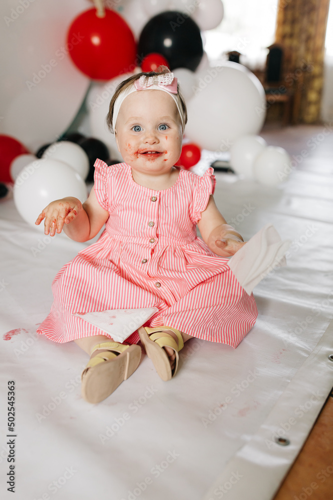 Fototapeta premium beautiful little one-year-old girl in a pink dress eats a lollipop. the child smeared himself with candy. kids love sweets