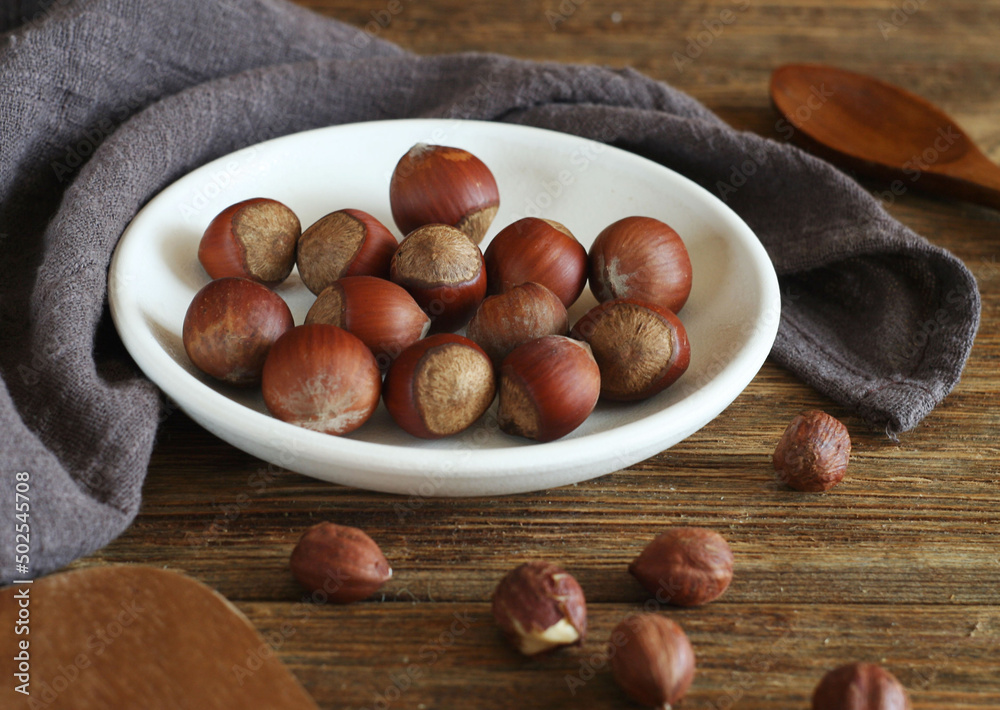 hazelnut lies in a bowl on a wooden table. Nearby lies a peeled hazelnut. Next to it is a cotton towel. hazelnut lies in a bowl on a wooden table. Nearby lies a peeled hazelnut. Next to it is a cotton towel.