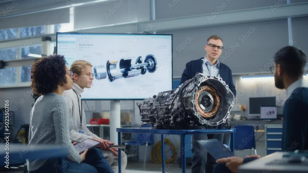 Male Chief Engineer Talk to Young Specialists About Components of the Electric Motor Using Projection Screen. People Ask Questions. Engineering and Green Electric Energy Concept