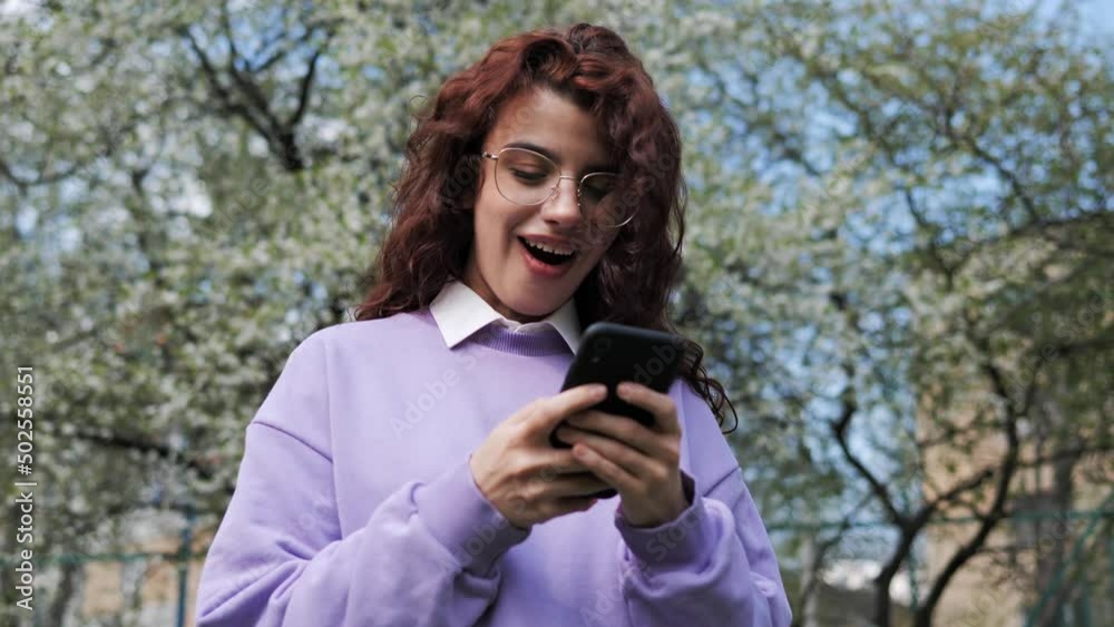 Joyful girl with curly red hair reading good news on mobile phone in slow motion. Overjoyed female freelancer celebrate online win success, read great news using smartphone, feel amazed outside. Joyful girl with curly red hair reading good news on mobile phone in slow motion. Overjoyed female freelancer celebrate online win success, read great news using smartphone, feel amazed outside.