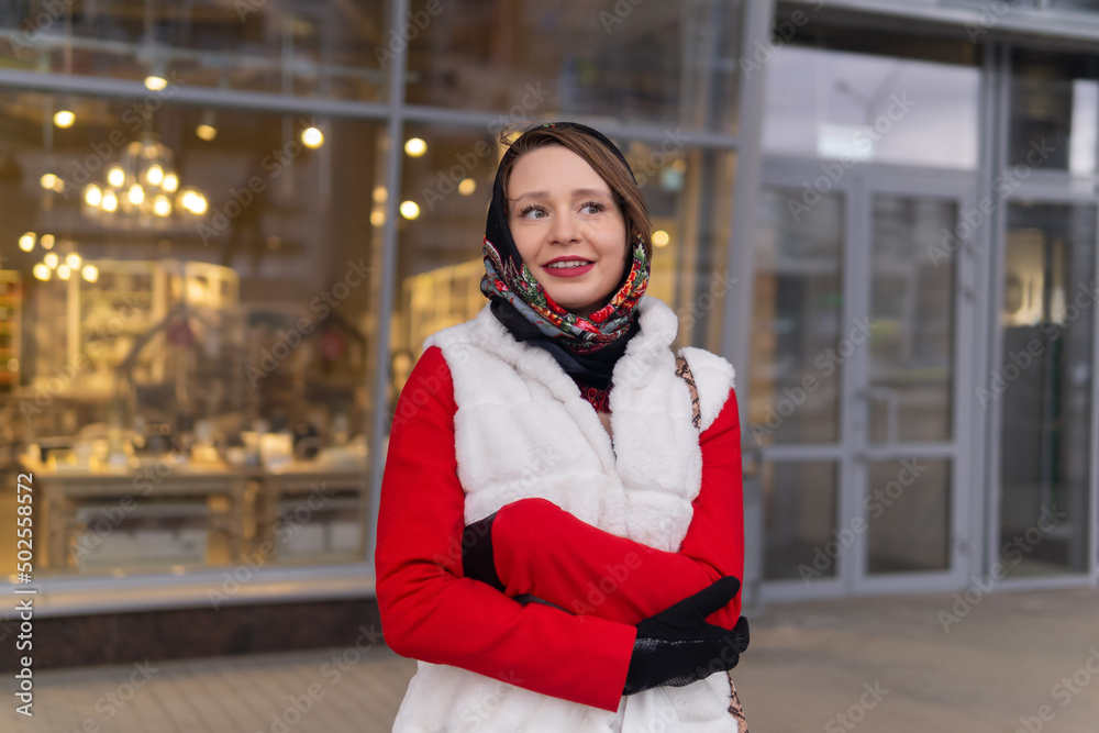 happy dreamy young woman in a headscarf in cold weather stands outside against the background of a blurry bright shop window