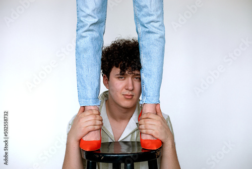 Portrait of young curly handsome man sitting at black stool holding feet of unrecognizable woman standing on stool.