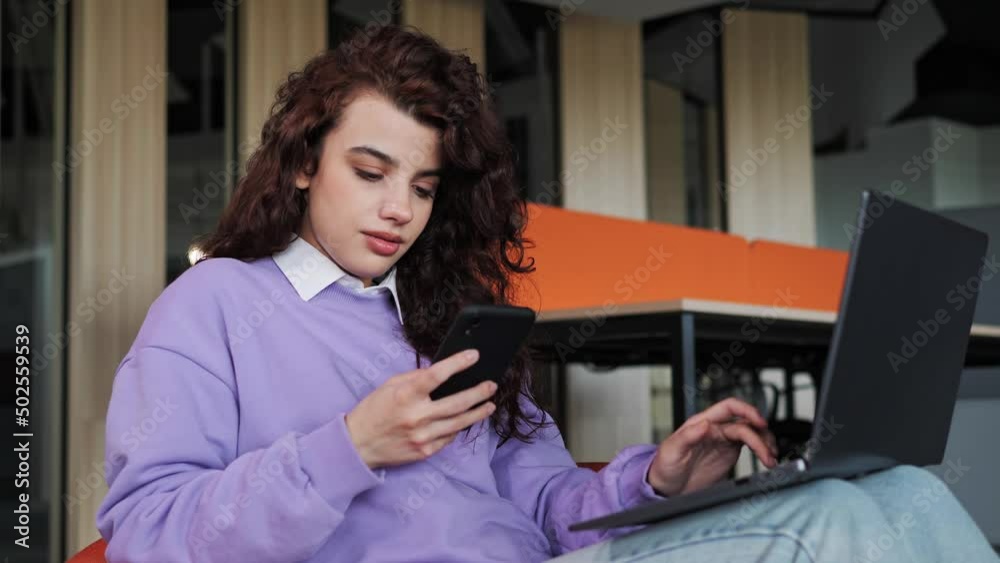Smiling young business woman professional using mobile phone working on laptop sits in comfortable chair typing message. Female customer make confirming online website shopping order delivery concept.