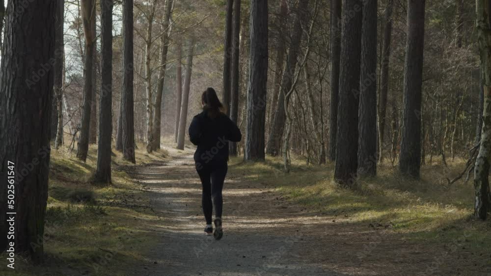 Fitness healthy jogger woman runs along forrest trail away from the camera towards background. Fit young woman with high tempo speed when running in the shade of trees on a sunny warm summer day.