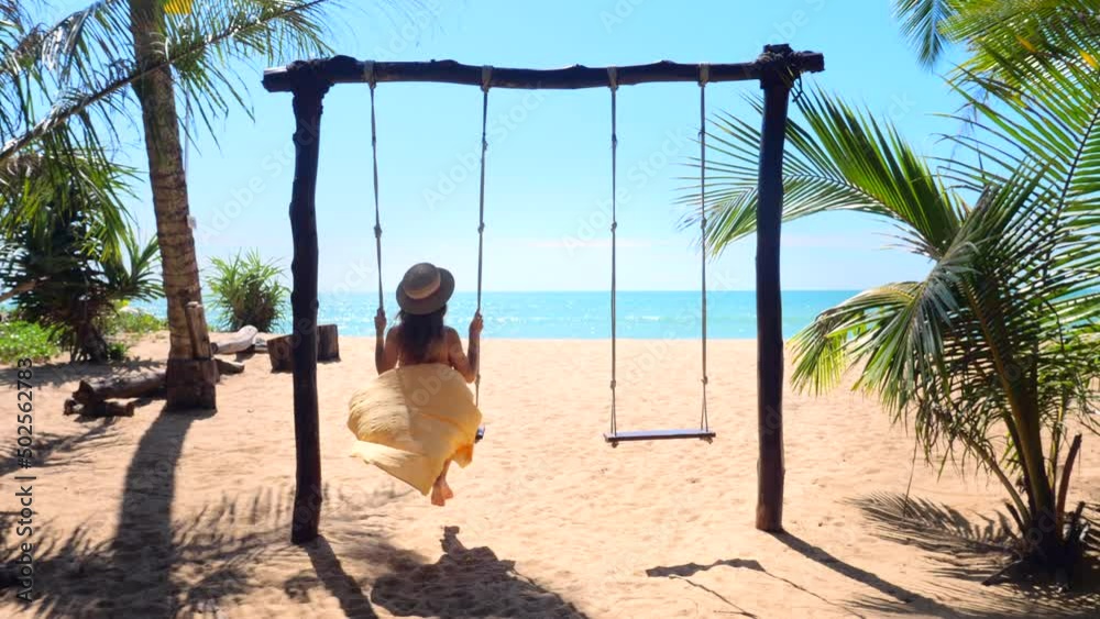 Woman with hat enjoying freedom on swing in Phuket, Thailand. Life, adventure and travel concept. Happy young woman in long dress on swing enjoy summer vacation in tropical resort with sea view